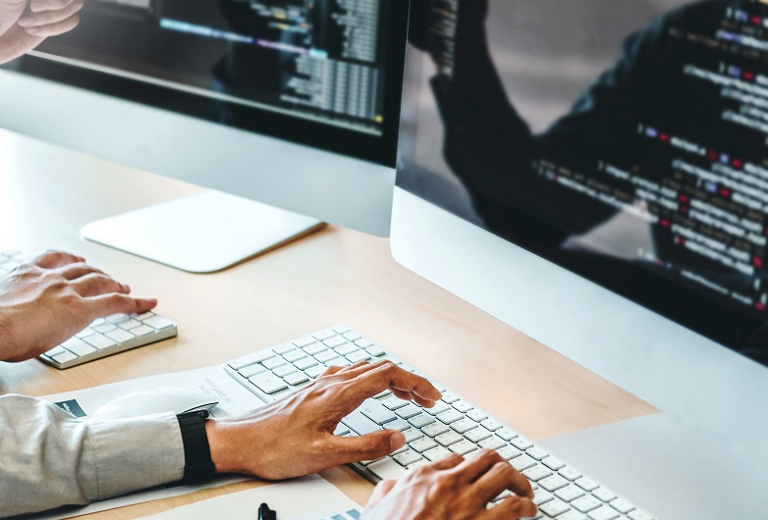 zwei Paar Hände tippen auf Tastaturen vor zwei mit Text gefüllten Monitoren